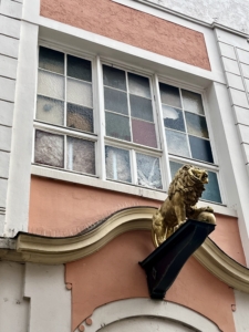 Drei weiße Fenster, darunter eine goldene Löwenfigur, die auf einem holzähnlichen Podest sitzt, das aus der Wand herausragt.