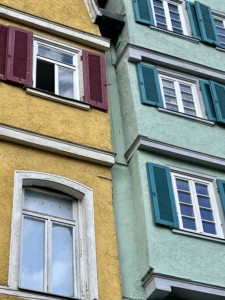 Ausschnitt von zwei nebeneinander stehenden Häusern. Das linke ist gelb mit weißen Fenster, die rote Fensterläden haben. Das rechte ist grünlich mit weißen Fenstern und blauen Fensterläden.