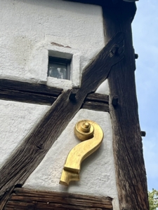 Kleines Glasfenster in einem Fachwerkhaus. Darunter befindet sich eine goldene Geigenschnecke, die an der Wand des Hauses angebracht ist.