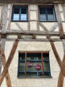 Drei Fenster mit blauem Rahmen in einem sandfarbenem Haus im Fachwerkbaustil. Auf dem unteren Fenster sind magentafarbene Sticker angebracht mit der Aufschrift "Eintritt frei".