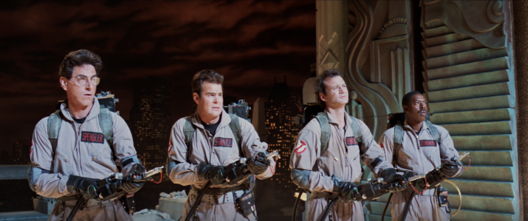 Der Screenshot aus dem ersten Ghostbustersfilm (Ghostbusters - die Geisterjäger (1984, USA) zeigt die vier Geisterjäger Geisterjäger Dr. Egon Spengler, Dr. Raymond „Ray“ Stantz, Dr. Peter Venkman und Winston Zeddemore. Sie haben braune Overalls an auf deren linken Brust ihre jeweiligen Nachnamen stehen. In ihrer hand halten sie den Protonenstrahler, der die Geister fangen kann.