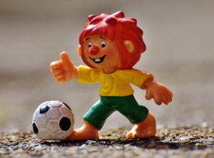 Zu sehen ist eine kleine Figur von dem Charakter Pumuckl, der seinen rechten Daumen in die Luft hebt und an dessen rechten Fuß ein Fußball liegt